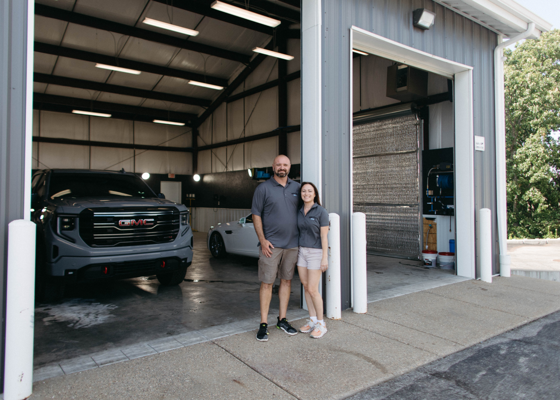 A man and woman stand outside a garage with vehicles inside.
