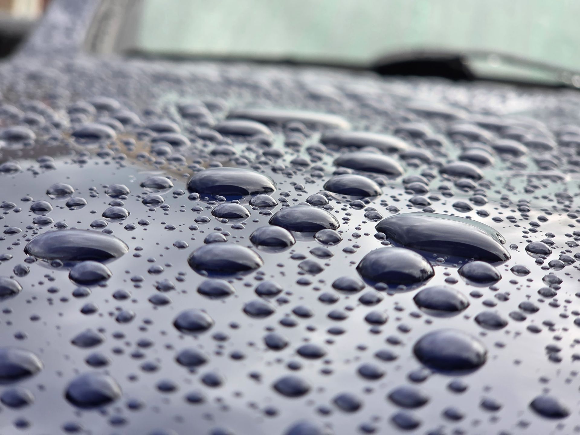Close-up of raindrops on a dark blue car hood, reflecting the sky.