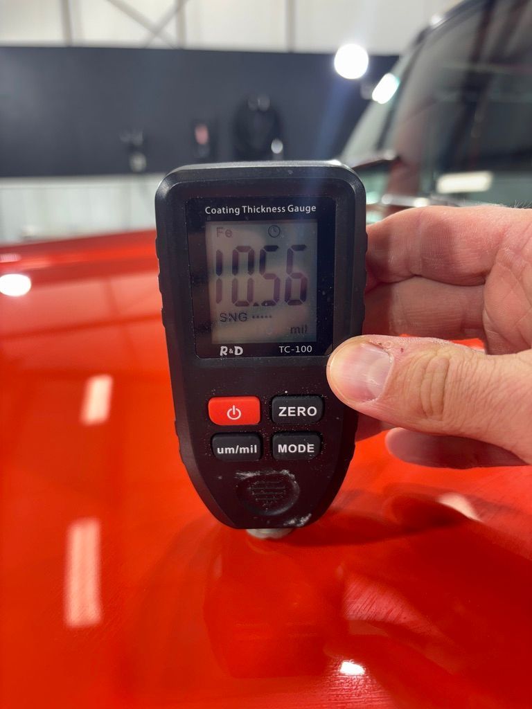 Hand holding a black paint thickness gauge on red car surface, displaying 10.56.