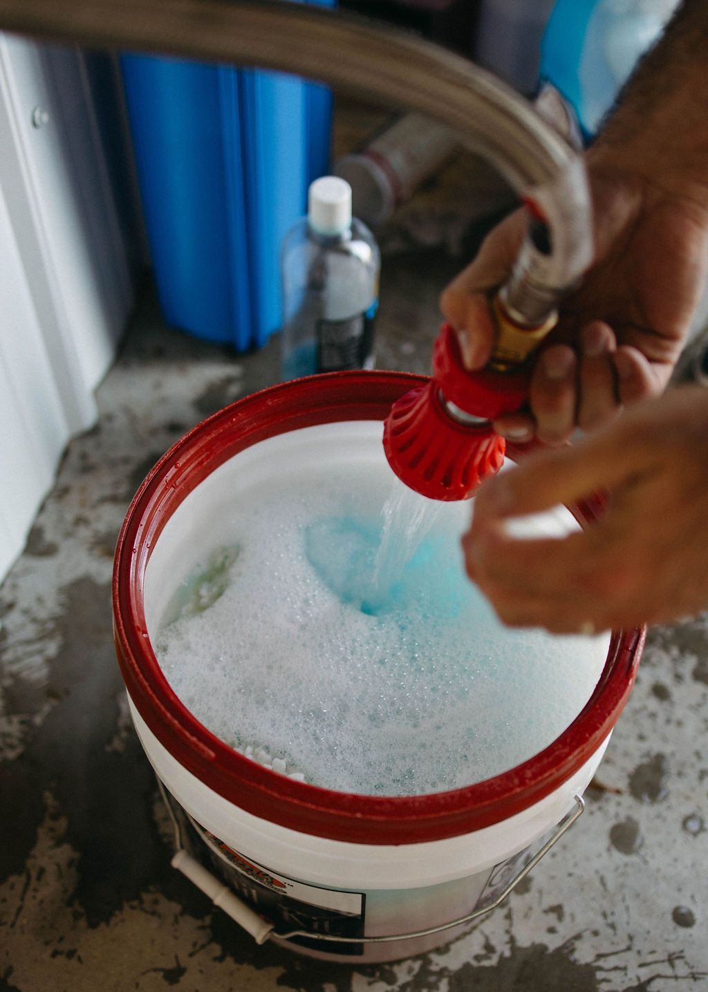 Person filling a red-rimmed bucket with soapy blue water from a hose nozzle outdoors.