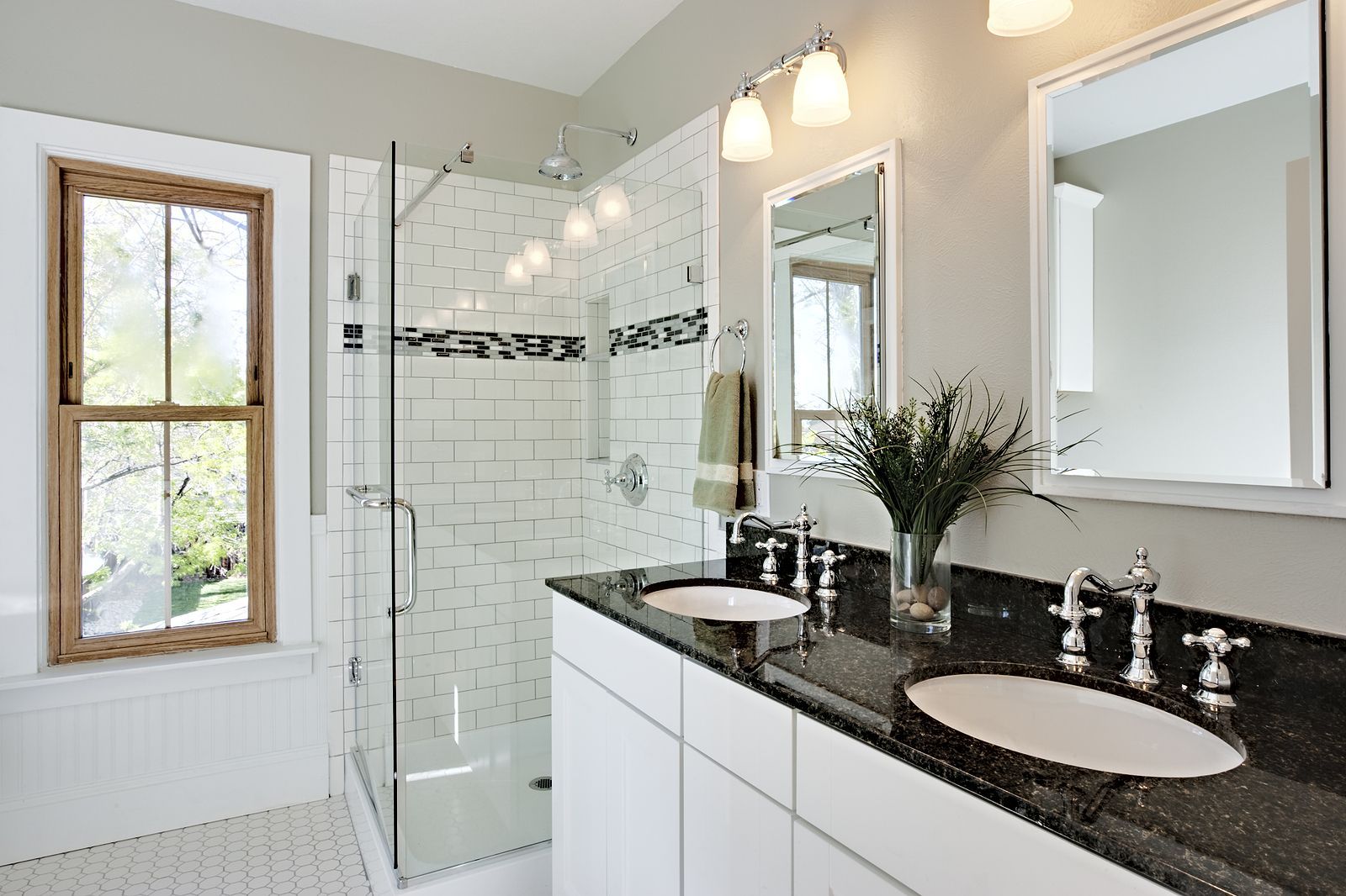Bathroom with a white tiled shower, double sink with black countertop, and large window.