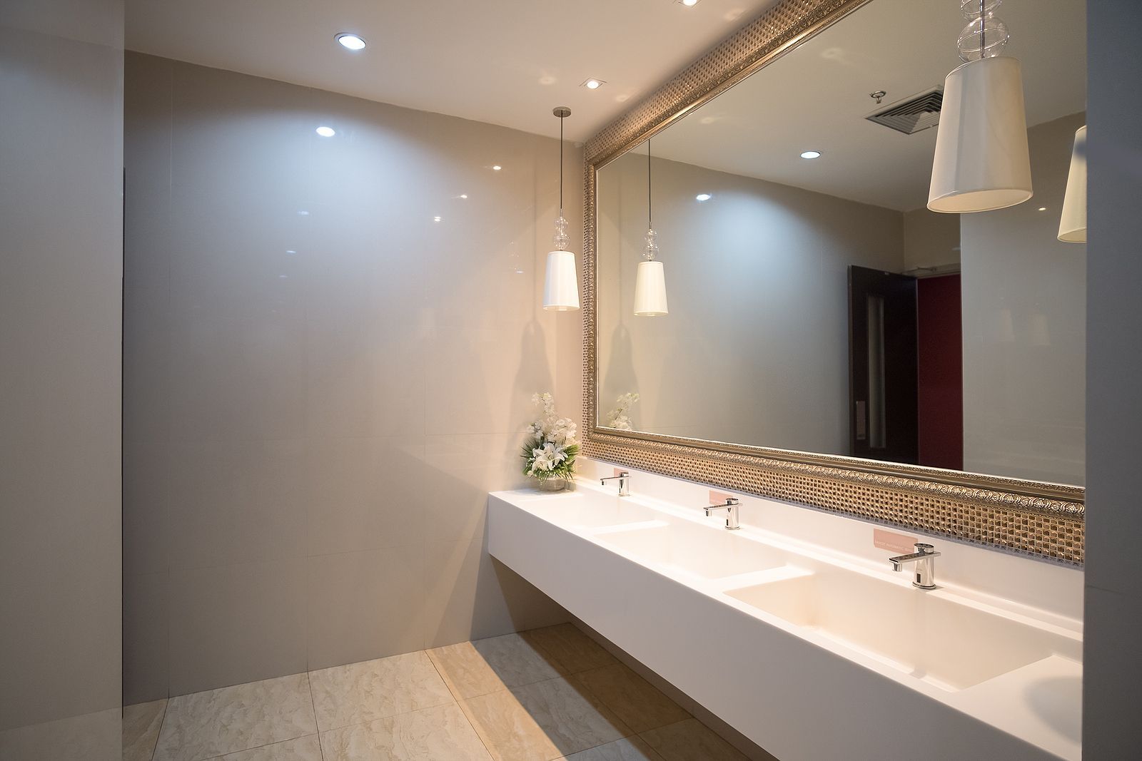 Modern restroom with a long white sink, large ornate mirror, pendant lights, and beige walls.