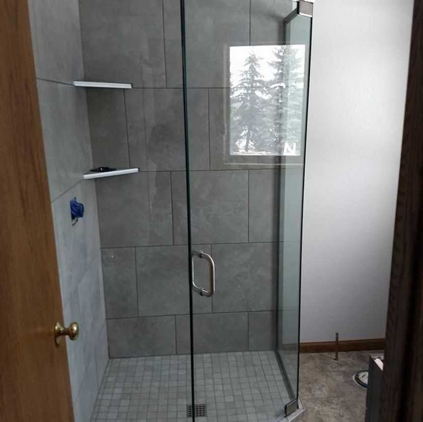 Shower with gray tiled walls, glass door, white shelves, and small window looking outside.