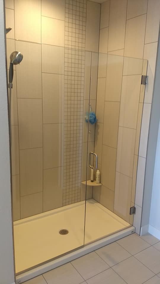 Glass-doored shower with beige tiled walls and a white shower base. Showerhead, soap, and loofah visible.