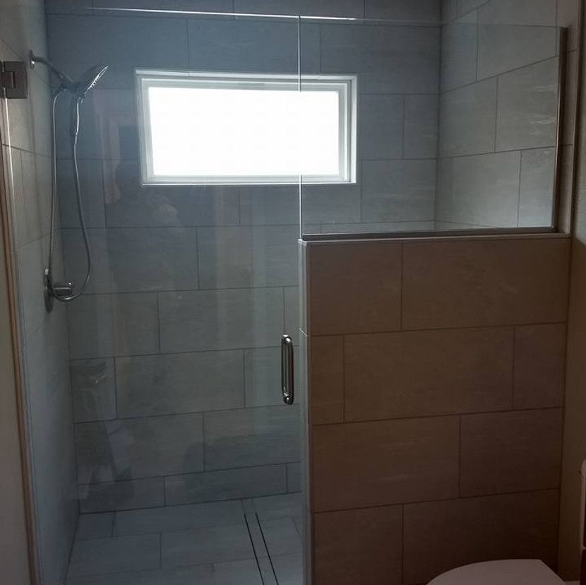Shower with glass door, gray tile walls and floor, window, and half wall.