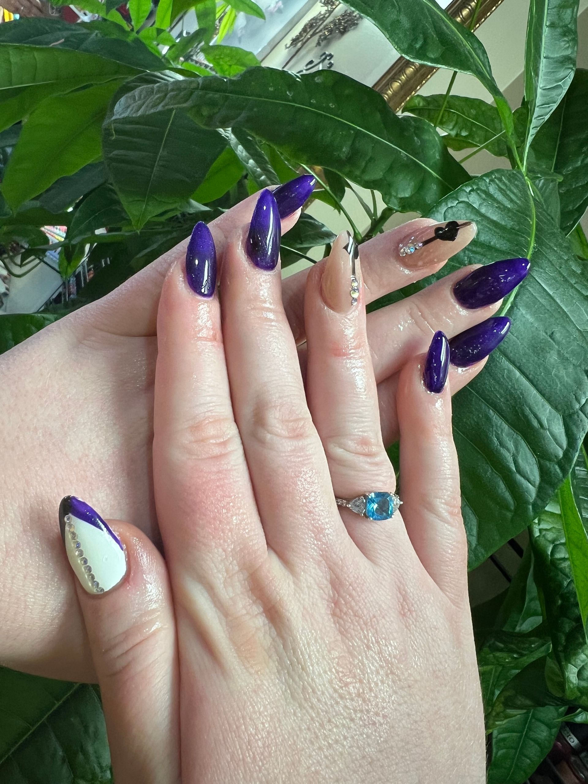 Hands with long, painted nails in a mix of purple, white, and neutral tones, decorated with glitter and designs, against a background of green plants.