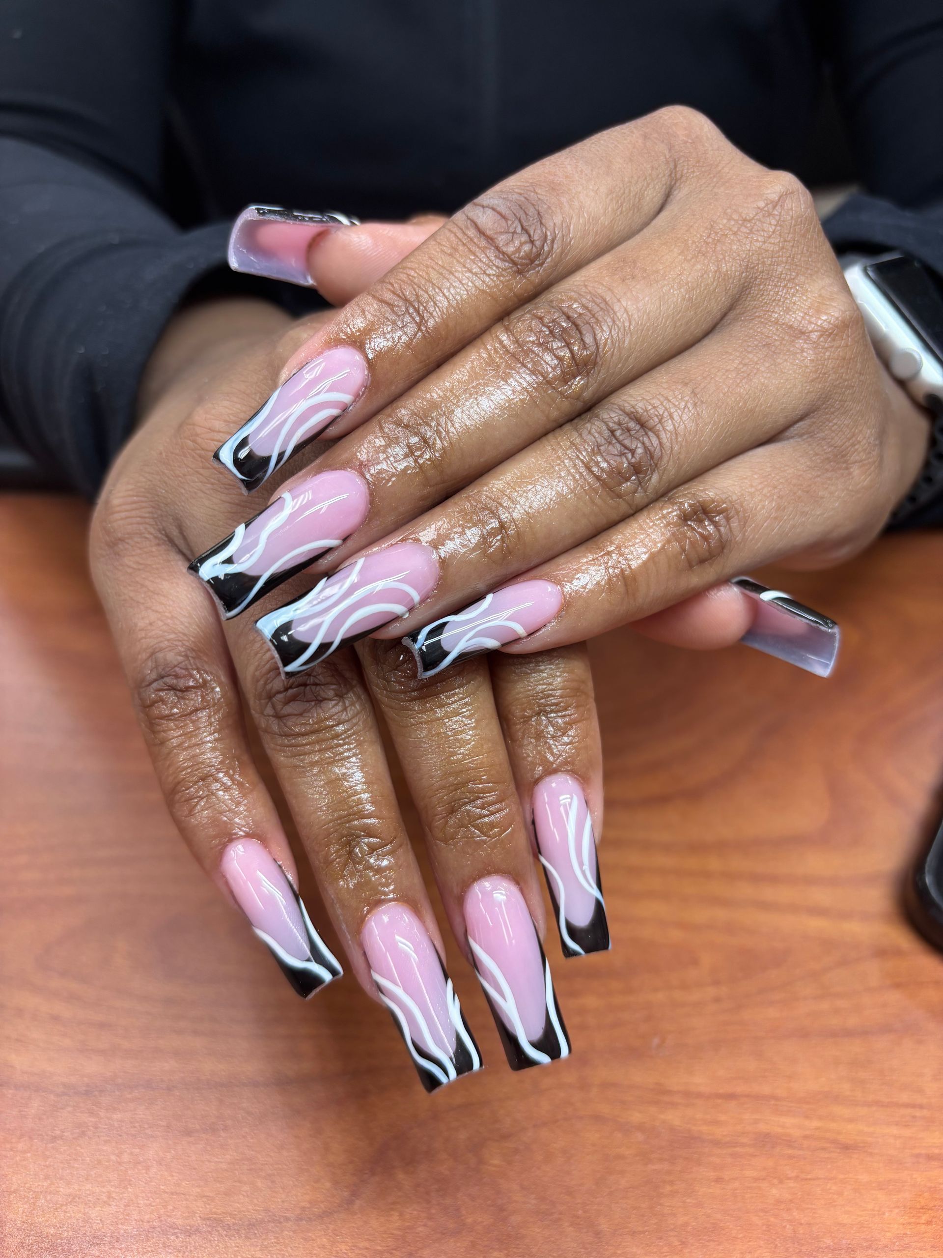 Hands with long, coffin-shaped acrylic nails. The design features a black French tip, white squiggle design, and pink base color.