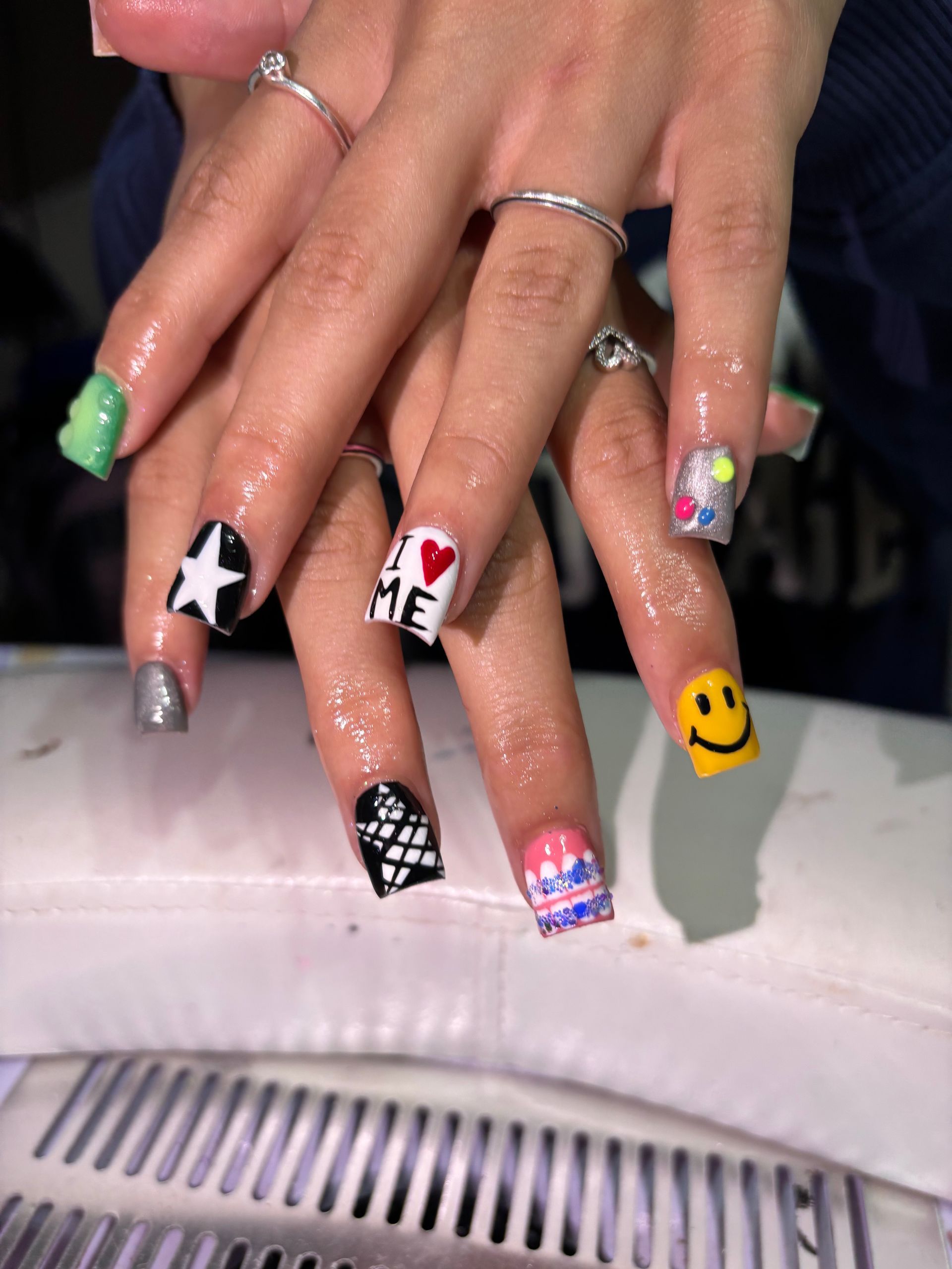 Hands with colorful, square-shaped acrylic nails. Designs include smiley faces, stars, hearts, and other patterns.