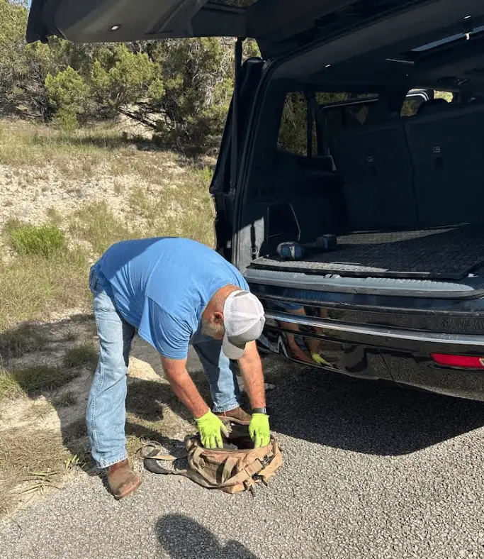 Man in blue shirt and jeans, wearing gloves, reaching into a bag next to a black SUV with its trunk open.