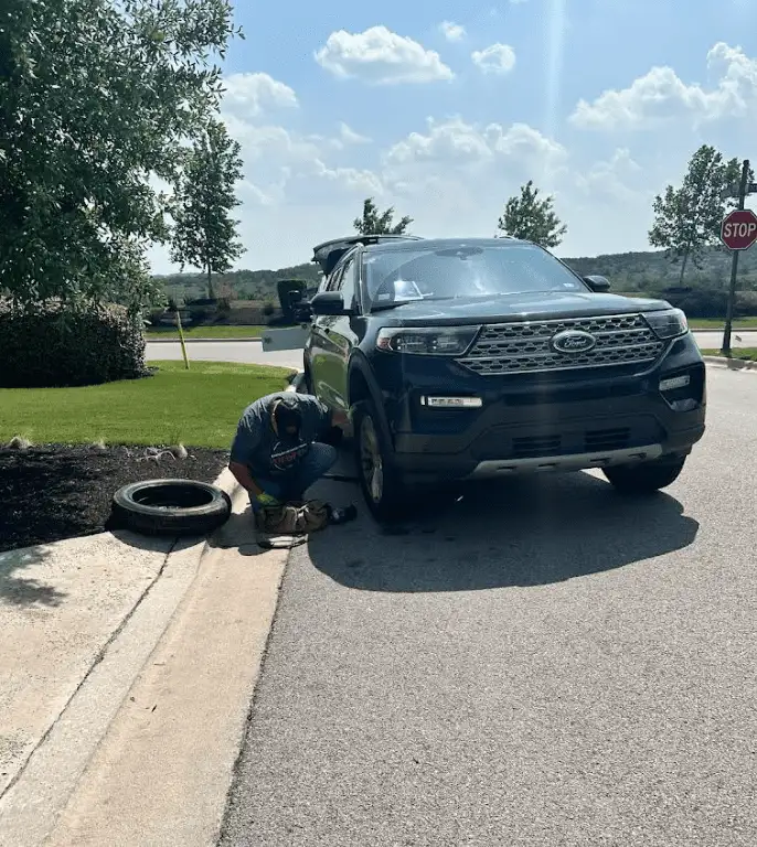 A person changing a tire on a black Ford SUV on a sunny residential street.