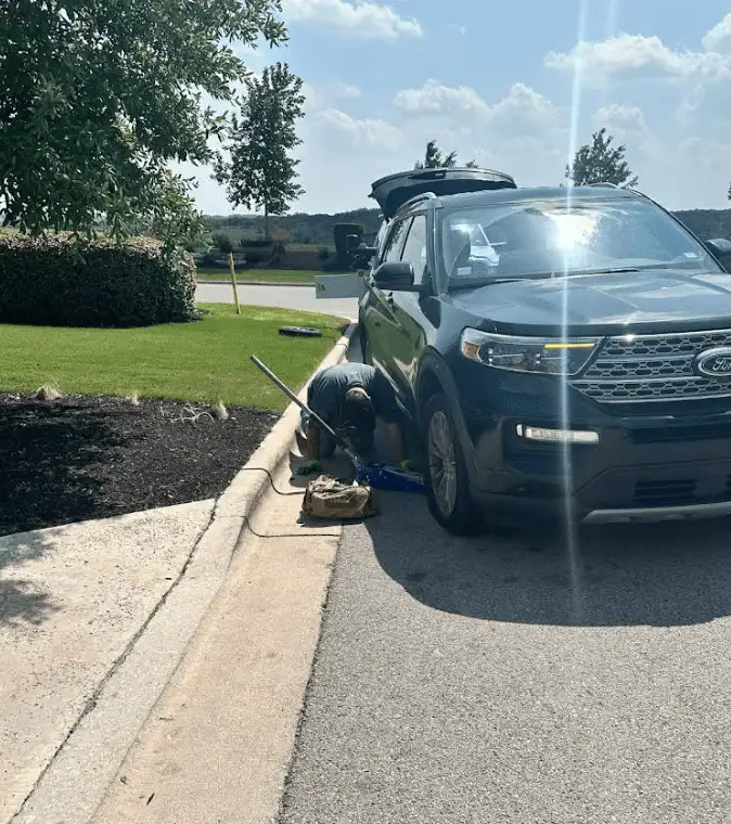 Person changing a tire on a black SUV parked on a curb in front of a manicured lawn on a sunny day.