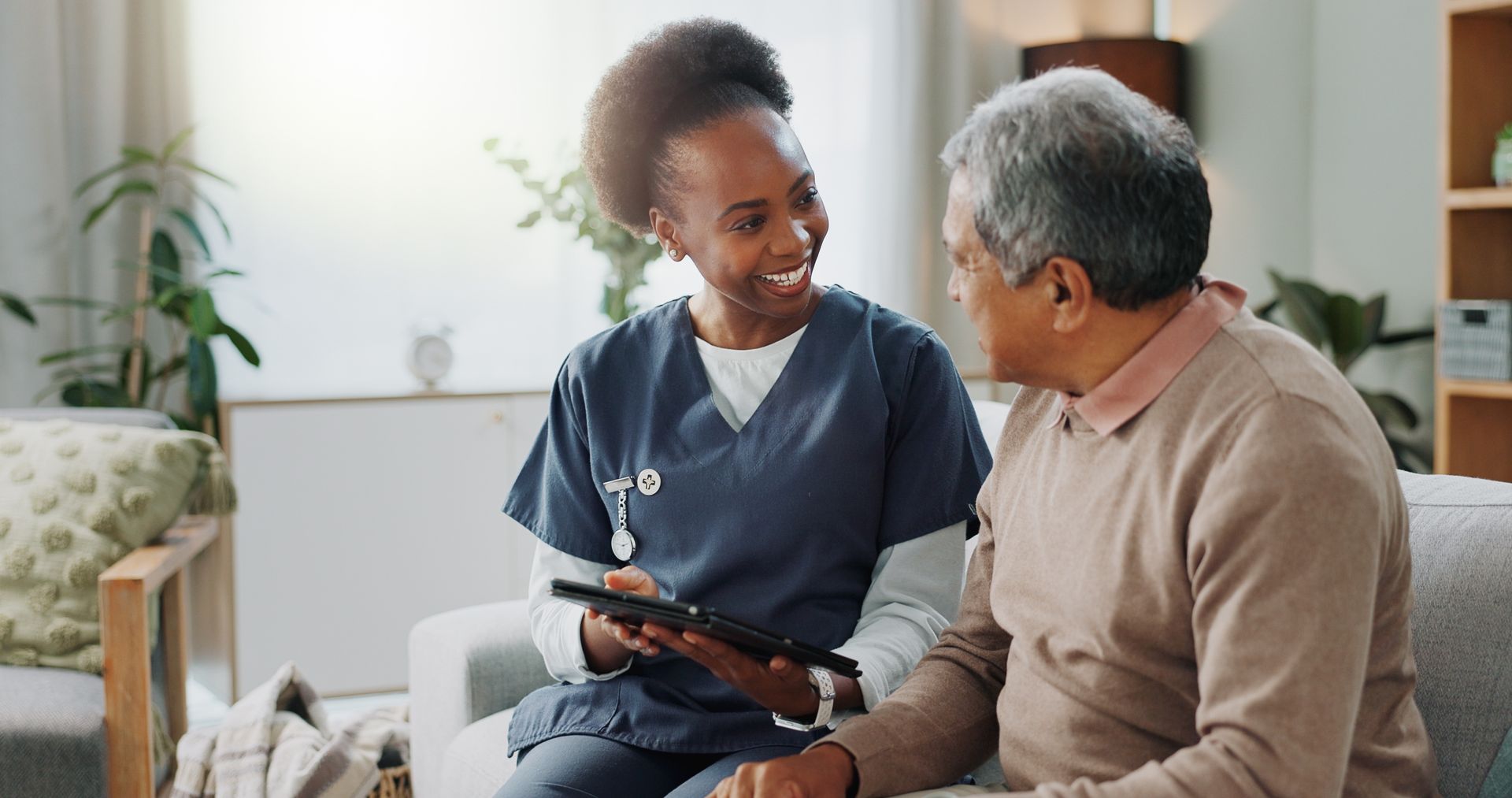 A nurse is sitting on a couch with an elderly man and looking at a tablet.