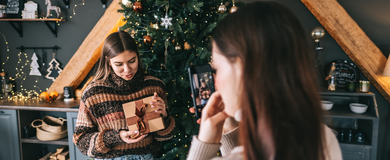 a woman taking a picture of another woman holding a gift in front of a christmas tree