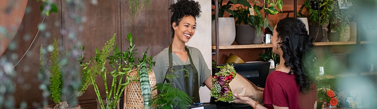 Two women exchanging flowers in a plant shop, surrounded by greenery and potted plants.