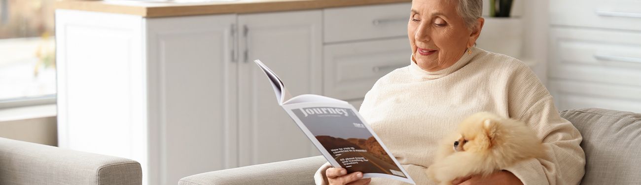A woman is sitting on a couch holding a small dog and reading a newsletter.