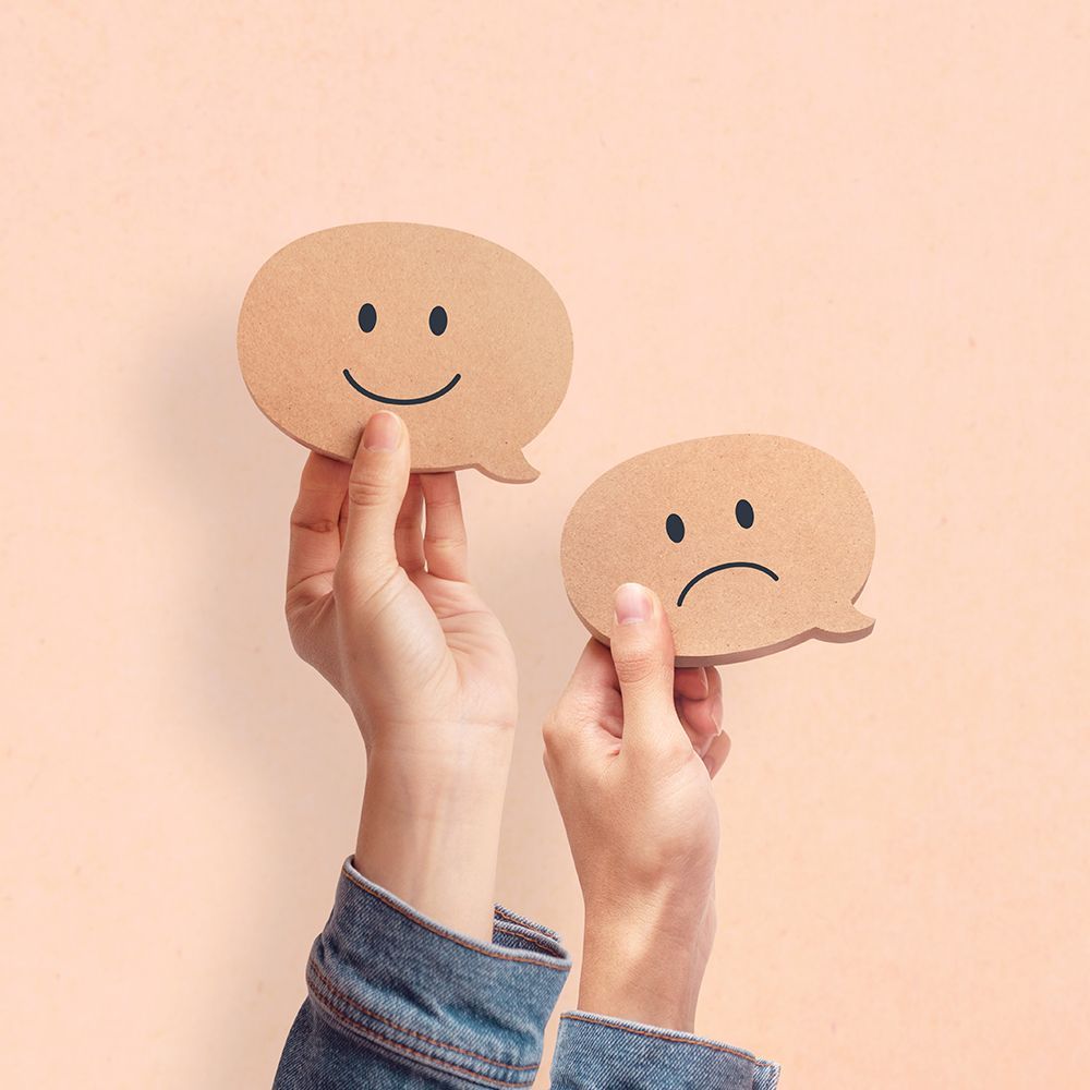 Hands holding two cardboard speech bubbles, one with a happy face, the other with a sad face.