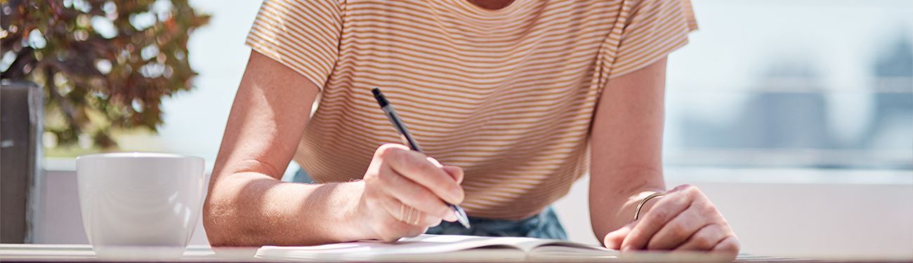 A woman is sitting at a table writing in a notebook with a pen.