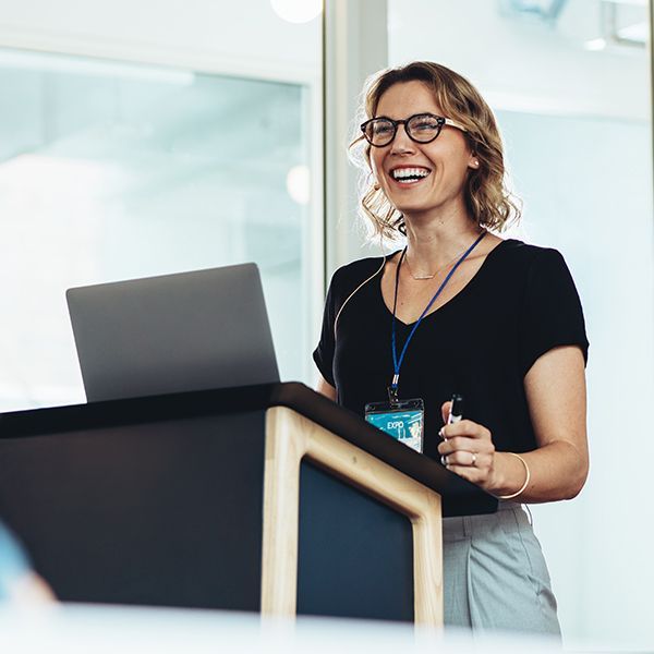 A woman is standing at a podium with a laptop and smiling.