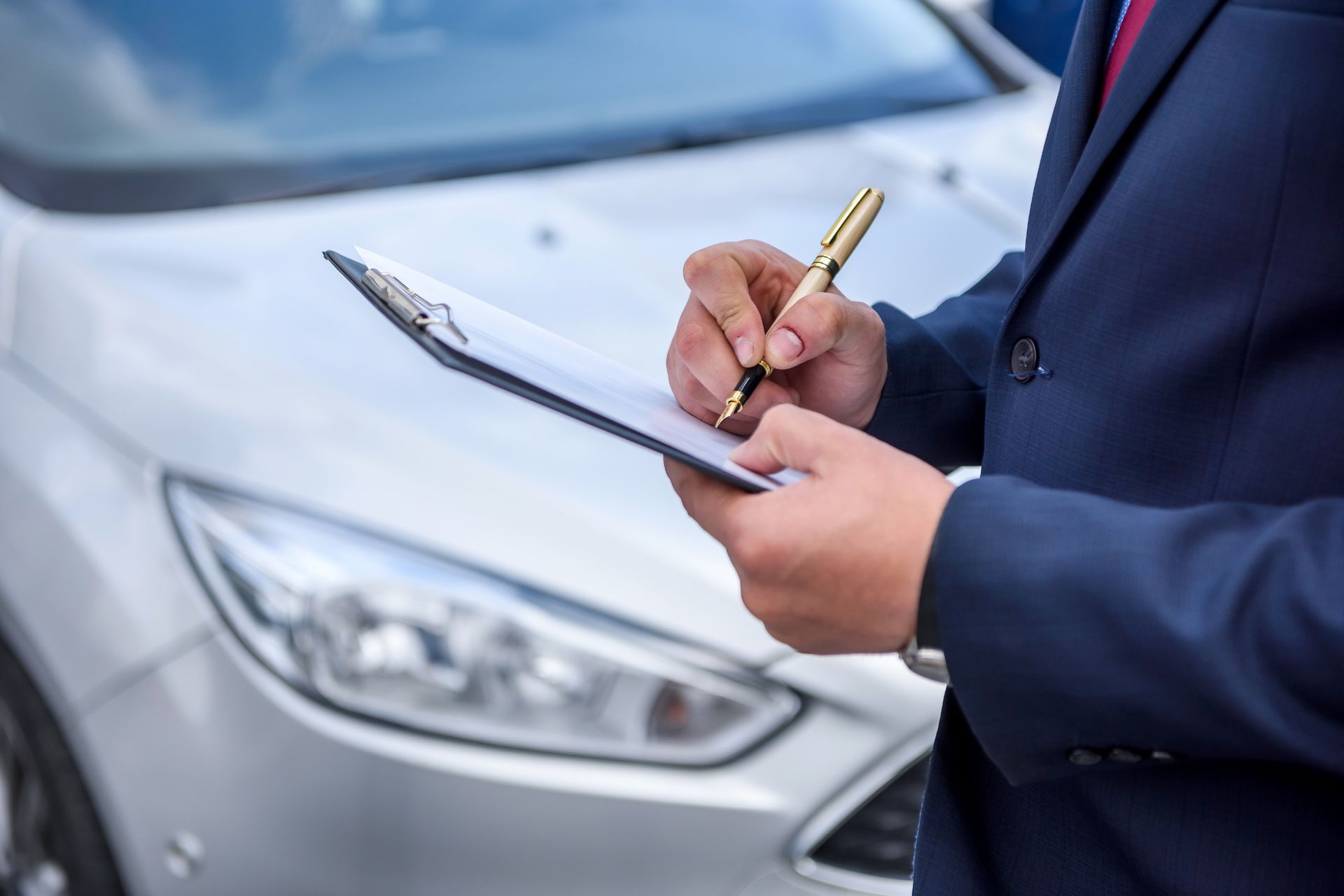 Person in suit writing on a clipboard, standing near a silver car.