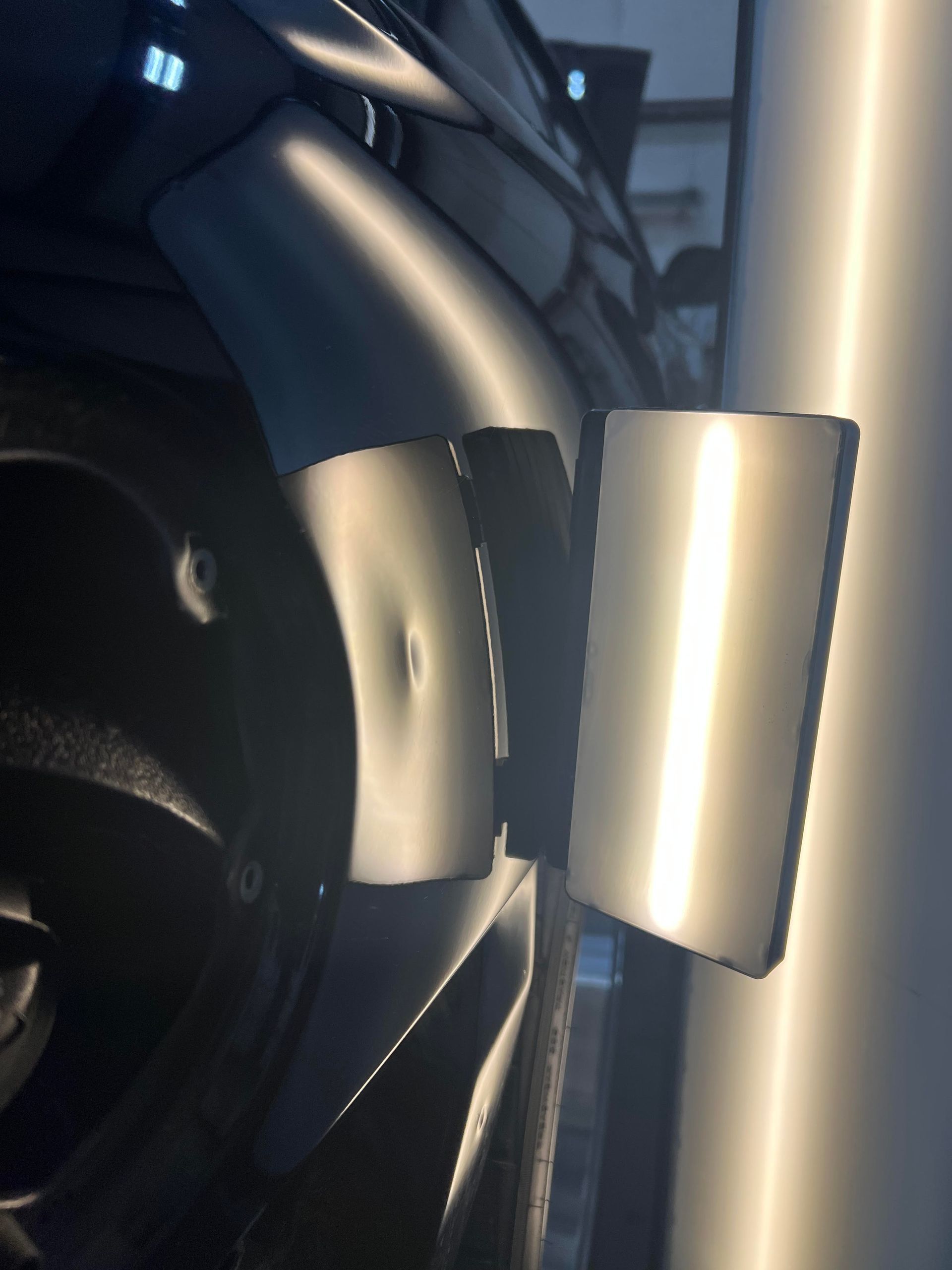 A small dent on a dark-colored car panel being inspected with a bright light reflection board.
