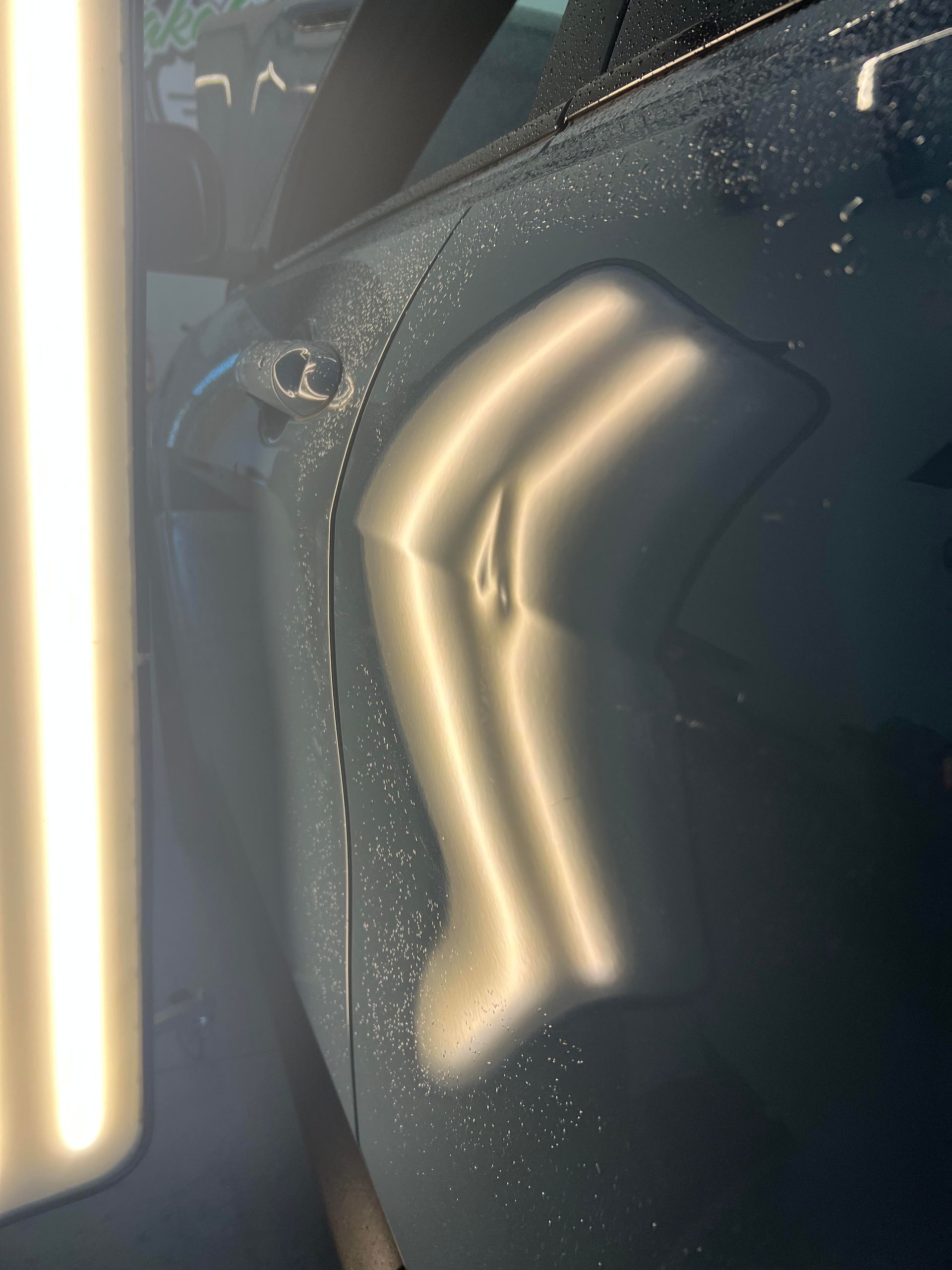 A close-up of a dented car door panel illuminated by a bright, vertical strip light for damage assessment.