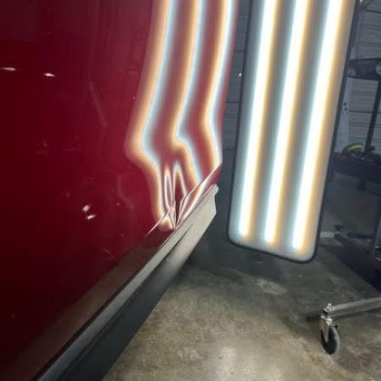 A close-up of a dented red car door being assessed with a light board to reveal surface imperfections.