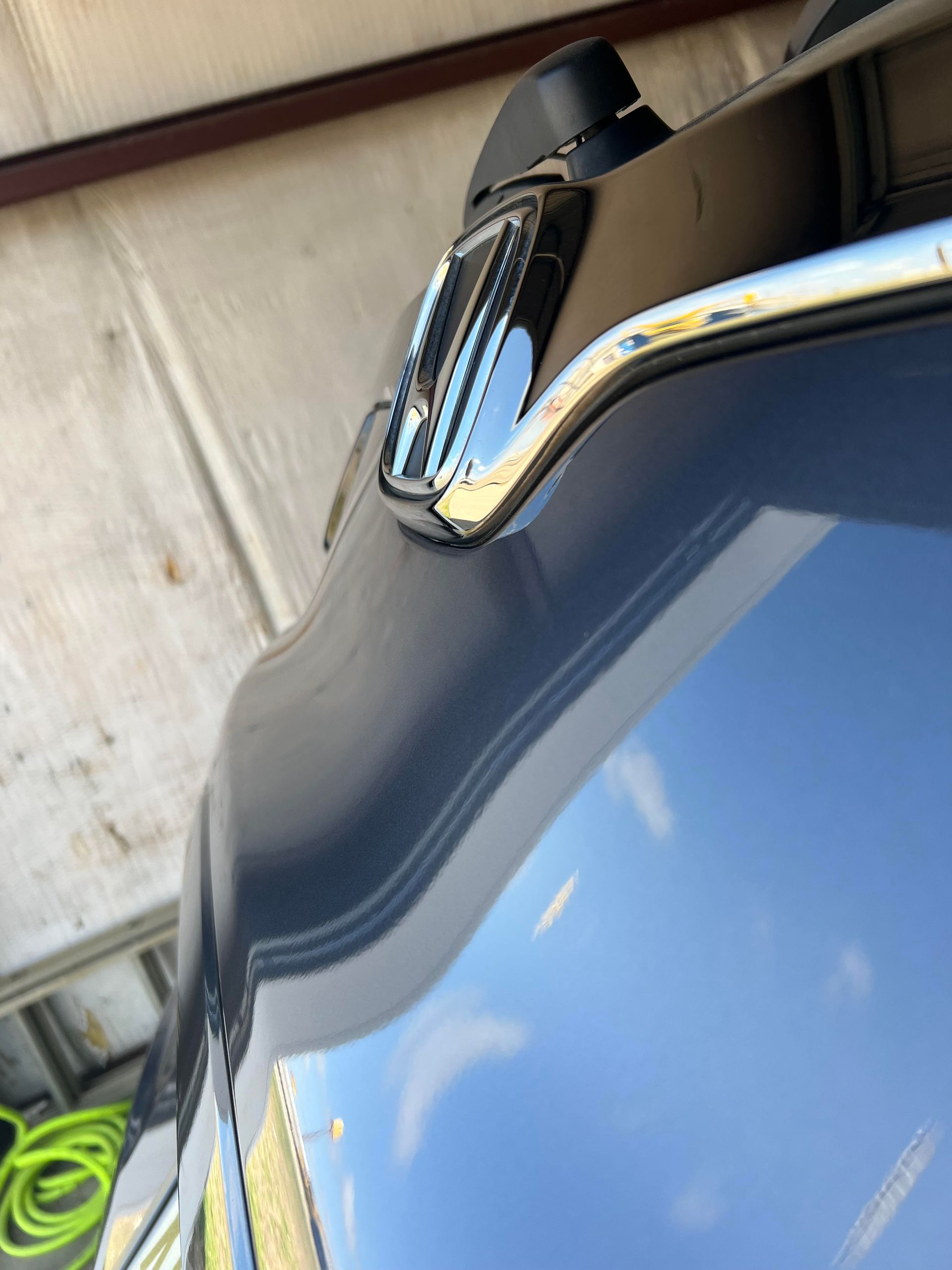 A close-up, angled view of a car's hood, chrome emblem, and trim, showing a blue vehicle reflected against a building.