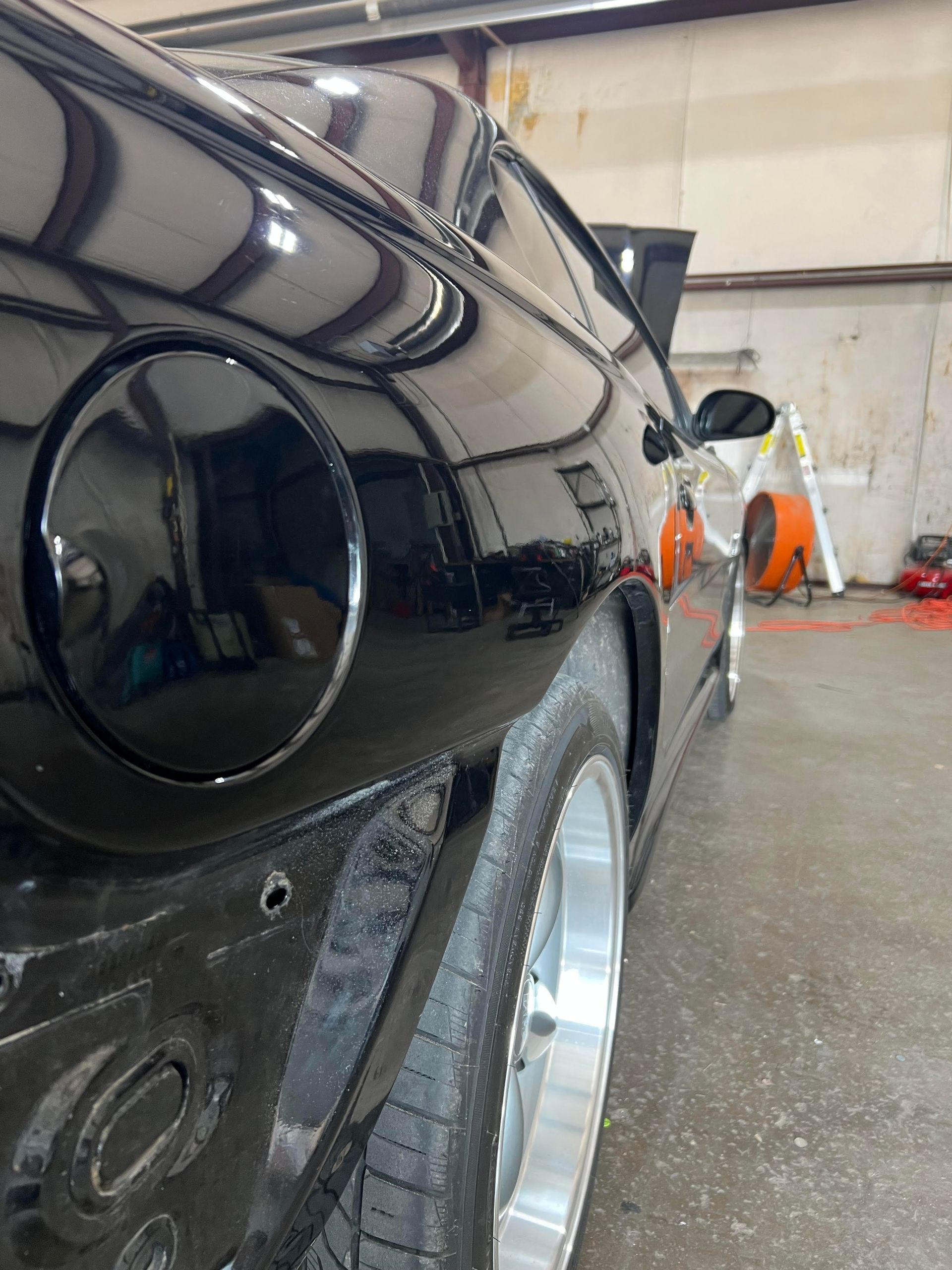 A close-up, angled view of a black car's rear quarter panel and tire in a garage, showing the fuel door and missing bumper.