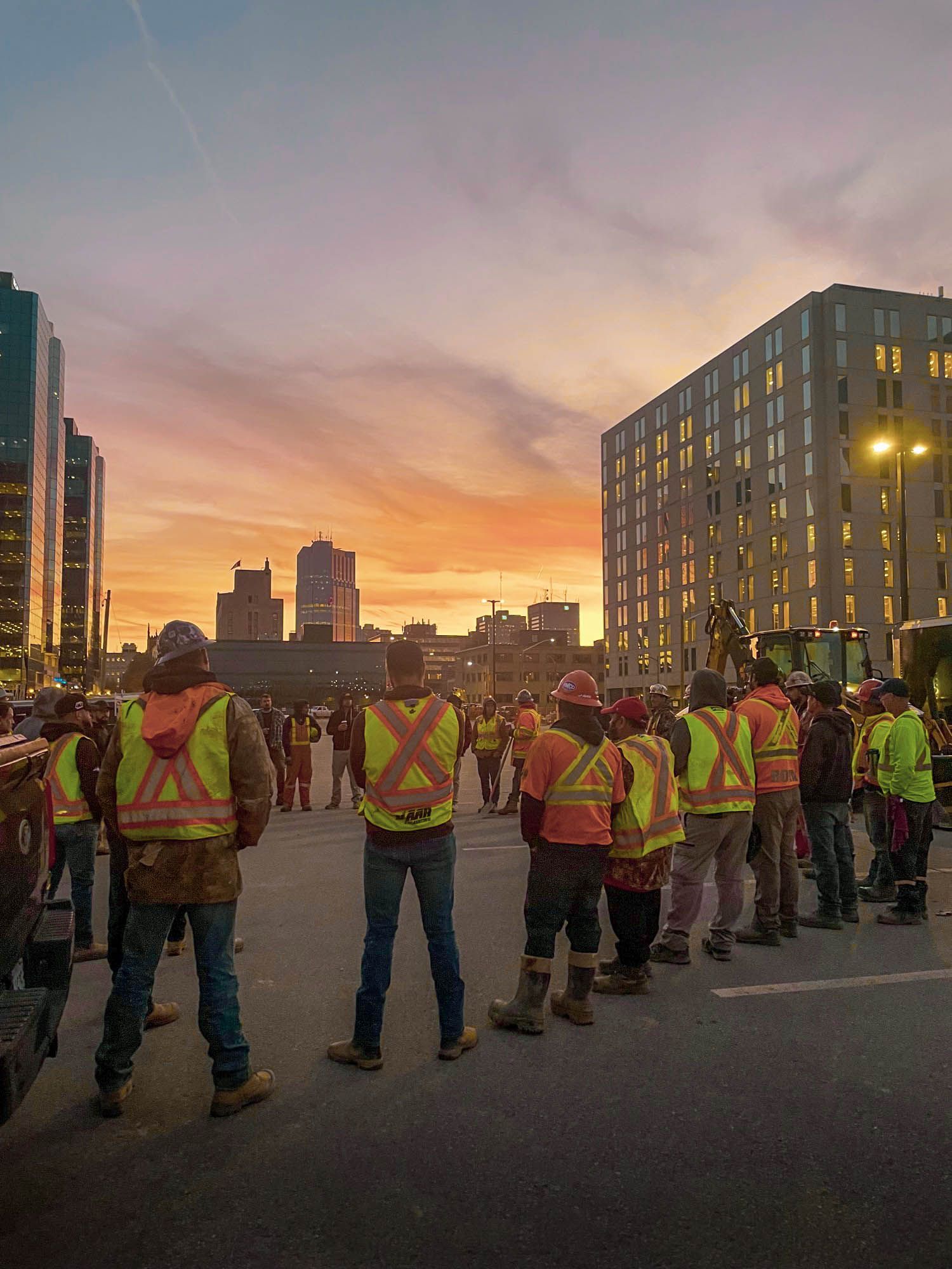 a group of construction workers are standing on a city street at sunset .