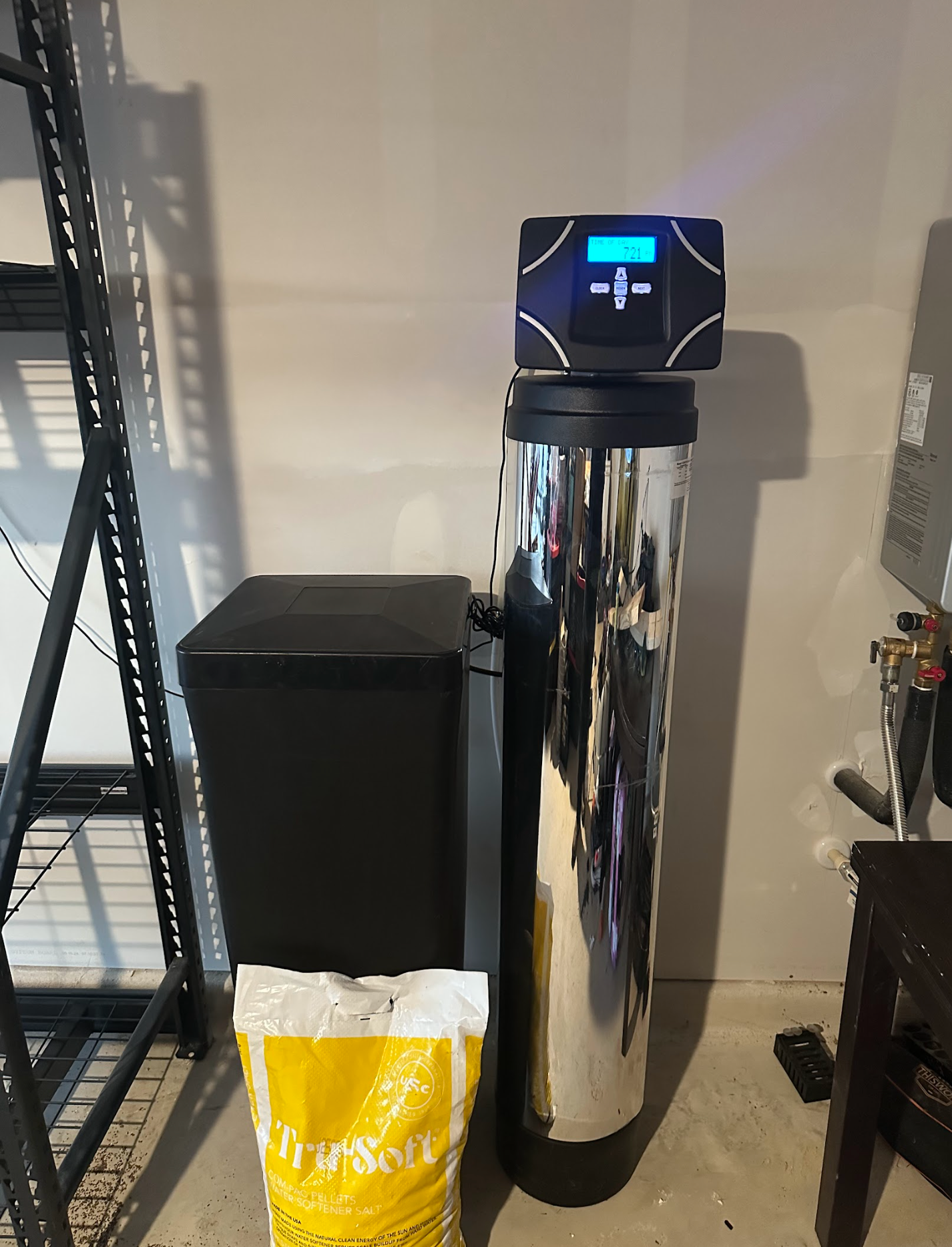 Water softener system in a garage, next to shelving and a salt bag.