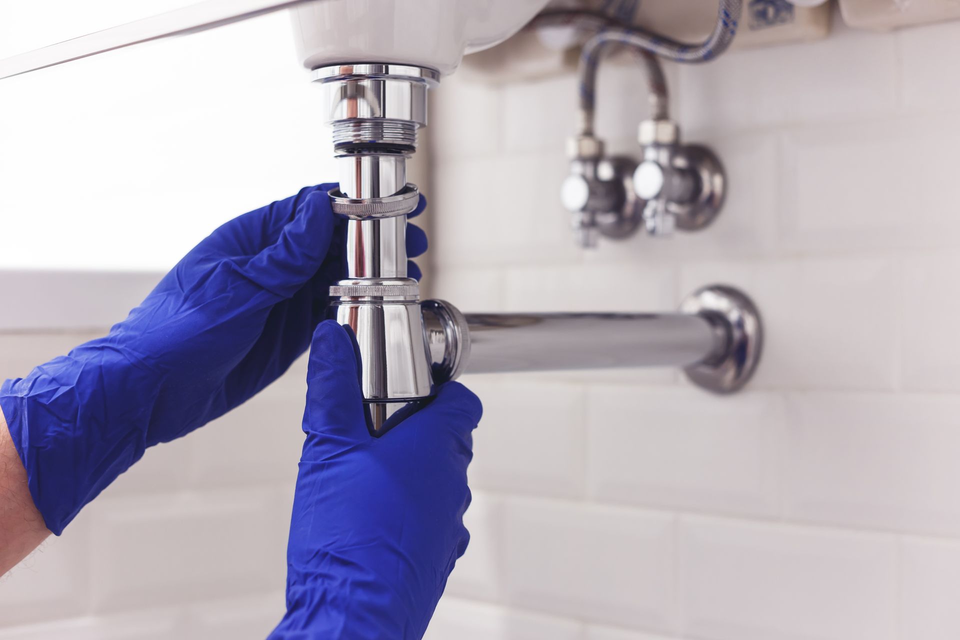 Hands in blue gloves, assembling a chrome plumbing trap beneath a white sink, in a tiled bathroom.