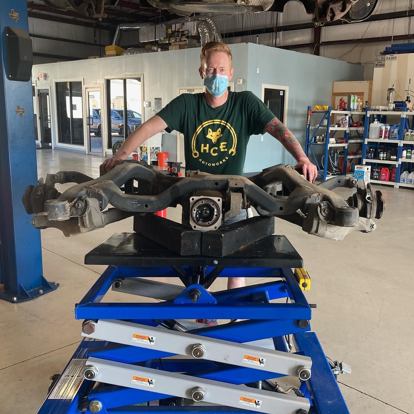 Man in mask holding large car suspension over blue lift in a garage. | Hill Country European Autoworks