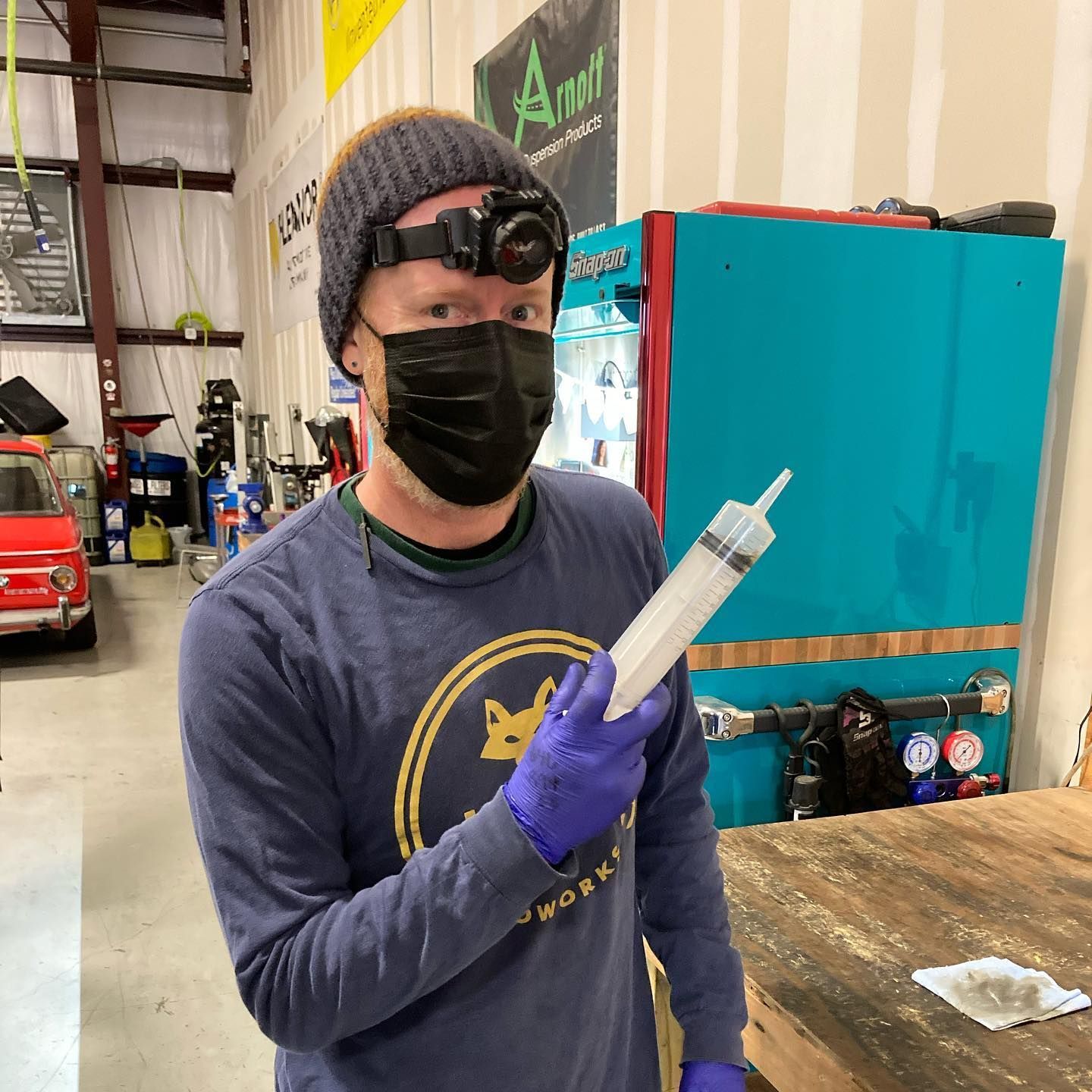 Man wearing a mask, holding large syringe in a workshop. He's wearing a beanie, gloves, and a headlamp. | Hill Country European Autoworks