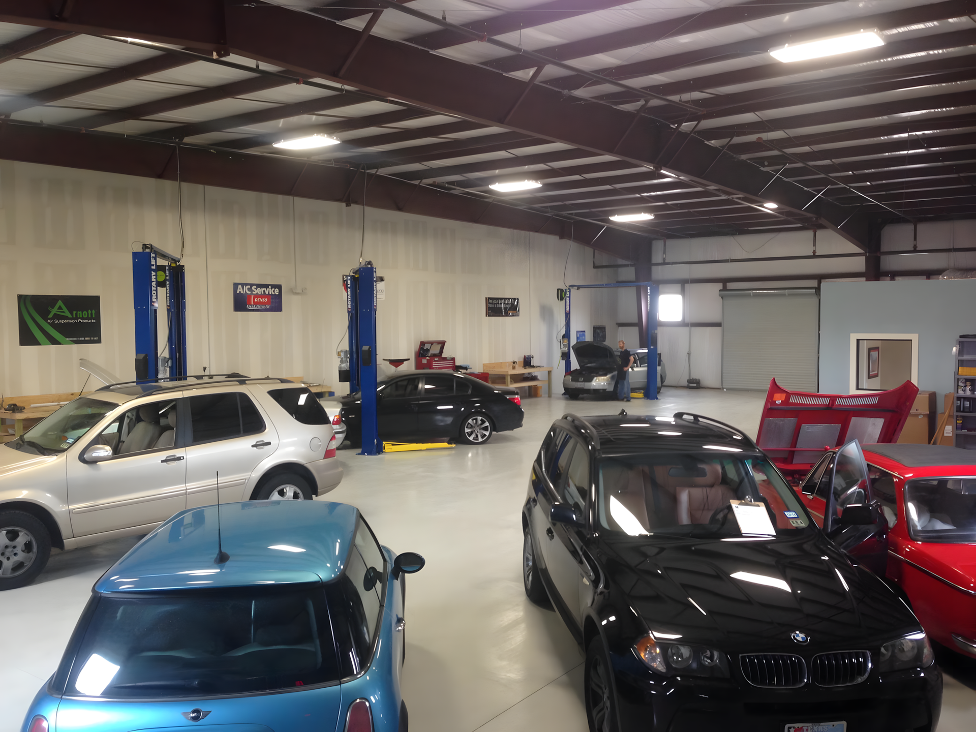 Cars in a well-lit auto repair shop, including a blue Mini Cooper and a black BMW, with lifts and tools. | Hill Country European Autoworks