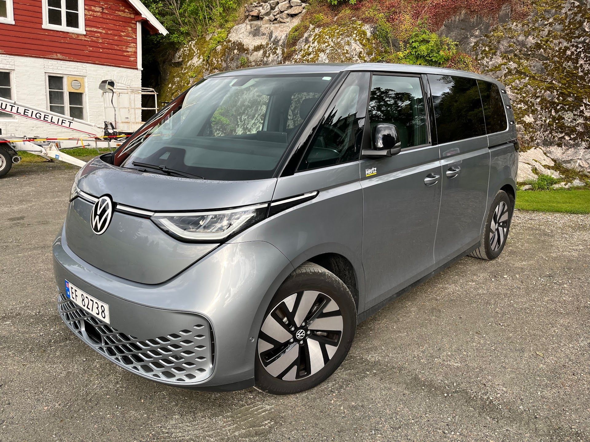 Gray Volkswagen ID. Buzz minivan parked on gravel, near a red house and trees. | Hill Country European Autoworks