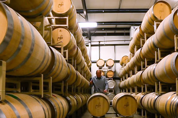 A man is standing in a warehouse filled with lots of wooden barrels.