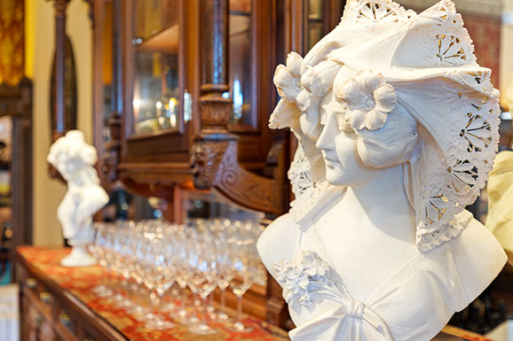 A statue of a woman is sitting on a table next to a row of wine glasses.