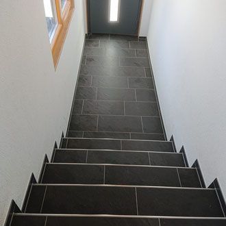 Treppe mit schwarz gefliesten Stufen und Wänden, die zu einer Tür mit einer darüber angebrachten Lampe führt.