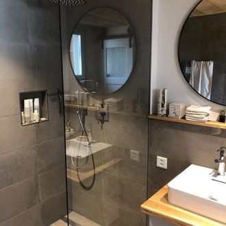 Modernes Badezimmer mit grau gefliesten Wänden, Glasdusche, runden Spiegeln und Holzregalen.