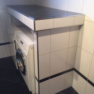 Eine Waschmaschine steht unter einer gefliesten Arbeitsplatte in einem Badezimmer mit weißen und schwarzen Fliesen.