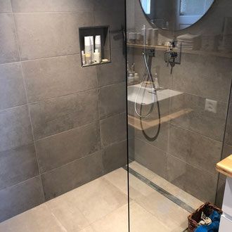 Modernes Badezimmer mit grau gefliesten Wänden, Duschtür aus Glas und eingebautem Regal.
