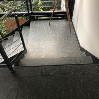 Treppe mit dunkelgrauen Steinstufen und Podest, Metallgeländer und einem Fenster mit Blick ins Grüne.