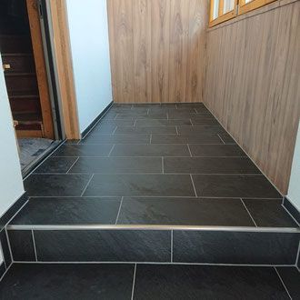 Eingangsbereich mit dunklem Fliesenboden, einer Stufe höher, holzgetäfelten Wänden und offener Tür links.