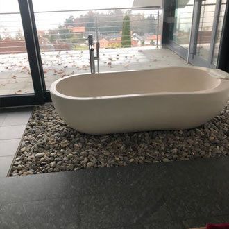 Weiße, ovale Badewanne auf einem Kieselbett, neben einem großen Fenster mit Blick ins Freie.