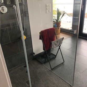 Dusche mit Glaswänden. Ein Handtuchhalter hält rote und graue Handtücher. Dunkel gefliester Boden.