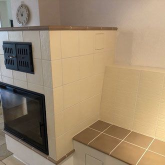 Kachelofen mit Glastür, Sitzbank und kleiner Tür, in Creme- und Brauntönen.