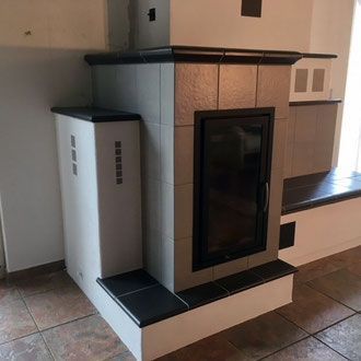 Gefliester Kamin mit schwarzer Tür, grauen Fliesen und weißem Sockel, in einem Raum mit Fenstersitzbank.