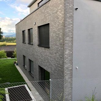 Modernes graues Backstein- und Stuckhaus mit dunklen Fensterläden, umgeben von grünem Rasen und einem Maschendrahtzaun.