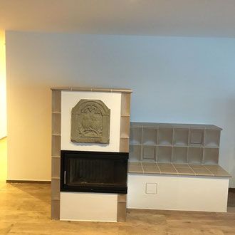 Gefliester Kamin mit dekorativer Schnitzerei und Bank vor einer hellen Wand.