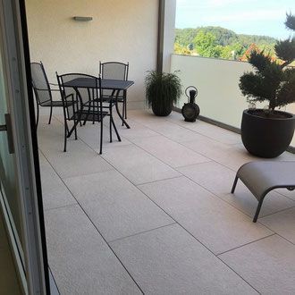Balkon mit Tisch, Stühlen, Pflanzen und Fliesenboden; im Hintergrund Blick auf Bäume.